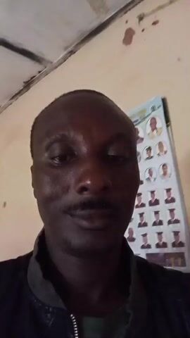 The media file is a close-up selfie video of a Black man with a shaved head and a mustache. He is wearing a dark jacket over a green shirt. He smiles and speaks briefly in the video. In the background, there is a poster on the wall with multiple small portraits of people. The room appears to be indoors with a textured ceiling and light-colored walls. The lighting suggests it is daytime. The setting appears to be a simple indoor space, possibly an office or classroom, given the poster. The man's expression conveys a friendly and approachable demeanor. The location context provided is Gure Kahugu, Nigeria.