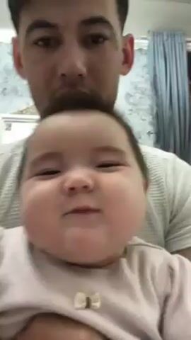 A man is holding a baby close to the camera. The baby is wearing a light pink shirt and has dark hair. The man is wearing a white t-shirt. The background shows light blue curtains and a picture frame. The baby is babbling and making noises. The man is looking at the camera.