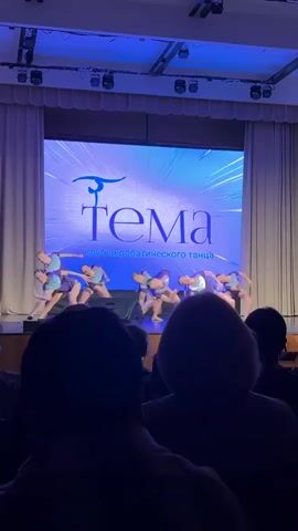 A group of young female dancers perform on stage in front of an audience. They are wearing matching purple and blue outfits. Behind them is a large screen displaying the logo of the dance club "Tema", which includes a stylized figure. The stage is well-lit with blue stage lights. The audience is seated in the foreground, their faces mostly obscured by the low lighting and the focus on the stage. The overall atmosphere is that of a formal performance or competition.
