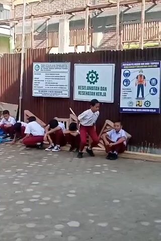 The video shows a group of young boys, likely students, dressed in white shirts and maroon pants, sitting on the ground in a line. They are positioned in front of a corrugated metal fence. The fence has several signs attached to it. One sign appears to be a safety poster with pictograms of people in safety gear and text in Indonesian. Another sign has a green cross symbol and text that reads "KESELAMATAN DAN KESEHATAN KERJA" (Safety and Health at Work). The boys are crouched down with their heads bowed, appearing to be in a moment of rest or instruction. One boy, standing at the end of the line, seems to be observing the others. The ground is paved with concrete. In the background, there is a building under construction, with scaffolding and building materials visible. To the left of the boys, there is a blue tarp. The overall scene suggests an outdoor school environment or a construction site where an educational activity related to safety is taking place.