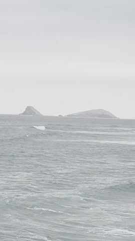 The video shows a static shot of the ocean under a cloudy sky. In the distance, two islands are visible, one to the left and one to the right. The waves in the foreground are white-capped and appear to be moving towards the viewer. The overall color palette is muted and gray.