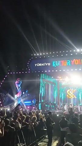 The video shows a live music performance at an outdoor venue at night. The crowd is dense, with people raising their hands. The stage is elaborately lit with multiple spotlights and a large LED screen displaying the performer. The screens also display "Türkiye Kültür Yolu Festivalli" and "T.C. Kültür ve Turizm Bakanlığı" at different points. The venue is crowded and the audience seems to be enjoying the performance.