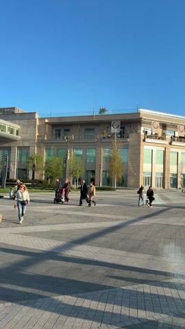 The video shows a sunny day at a modern outdoor shopping area or plaza. Several people are walking around, some in pairs, and others alone. There is a stroller in one group. The plaza is paved with large grey tiles. Behind the plaza are several modern buildings with large windows and beige exteriors. There is a body of water with a walkway in the background. The walkway has black metal railings and red flowers planted along it. The sky is clear and blue.