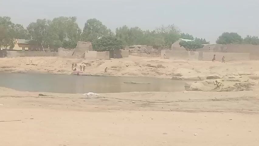 The video shows a small body of water surrounded by sandy banks. Several children are playing in and around the water. A group of people, including children, are sitting on the bank to the left of the frame.  In the background there is a sparse village with mud brick structures and trees. The day appears hazy.