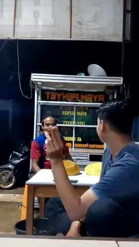 Two men are sitting at a small white table outside a food stall at night. The stall has the words "AYAM PENYET" printed in reverse on its front. One man, wearing a red and blue shirt, is seated opposite the camera, smoking a cigarette. The other man, wearing a blue shirt and black pants, is in the foreground and facing sideways. He is gesturing with his fingers as he speaks. Two yellow plates are on the table. A motorcycle is parked next to the stall. After a few seconds, the camera zooms out, showing more of the stall's structure and nearby trash cans. The lighting is dim.