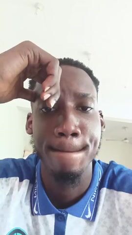 A medium shot shows a young Black man looking directly at the camera. He is wearing a white t-shirt with a blue collar. The collar has a design featuring the word "NIKE" repeated along it. He has short, dark hair and a small goatee. His left hand is raised, with his fingers touching his forehead, and he is making a facial expression that suggests mild irritation or contemplation. The background is a white ceiling with a few visible light fixtures.