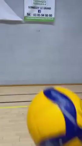The camera pans up from a polished basketball court with black and yellow lines to a basketball hoop mounted on a wooden support. The wall has several promotional signs, including "LES OPTICIENS GUILLON" and "CTF CONTROLE TECHNIQUE FERREIRA", with phone numbers. A yellow and blue volleyball is thrown from offscreen and bounces off the backboard and wall before bouncing off of the floor out of frame.