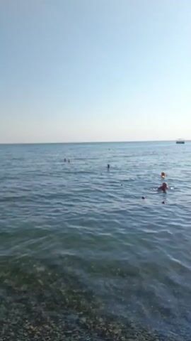 This media file captures a lively scene at a pebble beach on a sunny day. The clear blue sky and bright sunlight reflecting off the gentle waves suggest a warm, pleasant time for a beach outing. Several people are visible in the water, enjoying a swim and the cool sea. A young girl, wearing an orange floatation device, is seen wading near the shore, her silhouette highlighted by the sun. Further out in the sea, other individuals are swimming and playing. A boat is anchored in the distance, adding to the sense of a vacation or leisure activity. The background is dominated by a lush, green hillside that slopes down towards the beach, indicating a coastal landscape. The beach itself is covered in grey pebbles, with a strip of sand and a few beach umbrellas and loungers suggesting a developed recreational area. The overall atmosphere appears relaxed and joyful, typical of a summer day at the beach. No visible text is present in the media file. The location context provided is Dyurso, Russia, which aligns with the described coastal scenery.