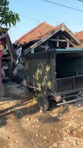 The video pans across a cluttered outdoor area, possibly a workshop or storage space, with a focus on a small truck and various items. In the foreground, a black truck bed is visible, its sides raised. To the left of the truck, a large blue gas cylinder stands upright. Behind the truck, a building with a damaged, open-roofed structure is seen. The interior of this building appears disorganized, with items like motorcycles, tools, and other miscellaneous objects scattered around. In the background, there are more structures with visible roofs and a clear sky. The ground is covered with dirt, debris, and various discarded items. The overall impression is one of disarray and potential disrepair.