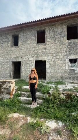 A woman with blonde hair walks up a set of stone stairs towards a dilapidated stone building. The building has open doorways and windows with no glass. The woman is wearing black leggings and a black sports bra. She is walking away from the camera. There is green vegetation growing around the stairs and the building. The sky is visible in the background.