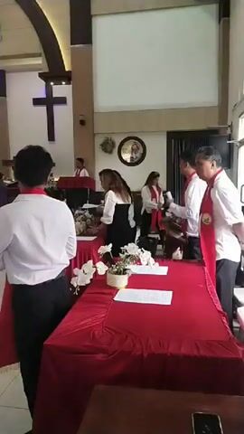 The video shows the interior of a church with a congregation gathered for a service. The scene begins with a view of the altar area, where several people in white shirts and red sashes are arranged. One person, facing away from the camera, wears a white shirt and black pants with a red sash. The altar is draped in a red cloth. The camera then pans to show rows of wooden pews occupied by people. Many are dressed casually, while some appear to be in more formal attire. The church features a cross prominently displayed on the wall behind the altar, and an alpha and omega symbol is visible above it. The lighting is bright, with ceiling lights illuminating the space. The overall atmosphere appears to be one of a religious gathering.