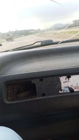 The video shows the interior of a vehicle, likely a car or van, from the perspective of the passenger seat. The dashboard is visible, with a cracked windshield. A photograph is stuck to the dashboard. Outside, there is a road with vehicles and some vegetation. The lighting suggests it is daytime, possibly overcast.