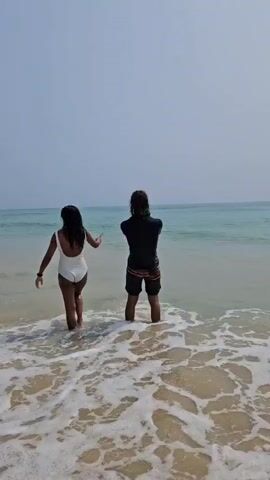 Two people are standing in the shallow water on a beach, facing the ocean. The person on the left is wearing a white one-piece swimsuit and has long dark hair. The person on the right has long hair and is wearing a black long-sleeved shirt and patterned shorts. The ocean is a light turquoise color, and the sky is a hazy blue. The waves are gentle, and the water is clear enough to see the sandy bottom.  The person on the right raises their phone to take a photo as the person on the left moves towards the water.