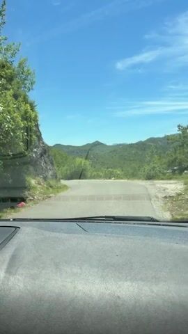 The video captures a view from inside a car, looking out through the windshield. The car is on a paved road that curves to the left, ascending a hill. The road is bordered by a steep, rocky embankment on the left and dense green vegetation on the right. The landscape is characterized by rolling hills covered in lush green trees, stretching towards the horizon under a clear blue sky with a few wispy white clouds. The dashboard of the car is visible in the foreground, with a black textured surface and a circular air vent. The lighting suggests it is daytime, likely a sunny day.