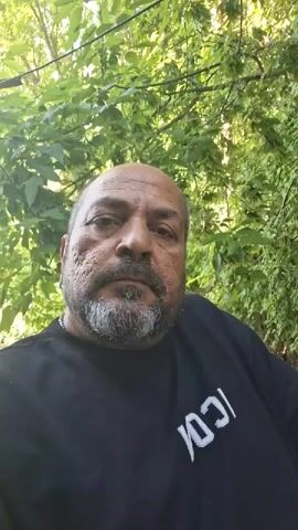 An older man with a bald head, a grey beard, and a mustache looks directly at the camera. He wears a black t-shirt with a white graphic that appears to read "ICON" backwards on the front. He has a serious expression and visible scarring on his left cheek. The background consists of lush green foliage and trees, indicating an outdoor setting. The lighting suggests daylight.