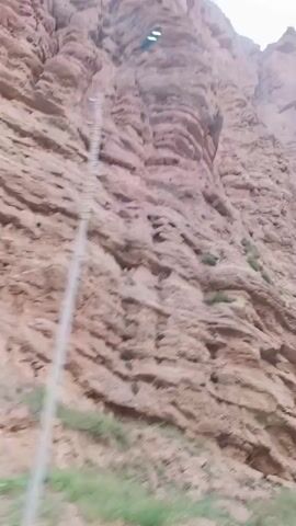 The video shows a rocky mountain with a reddish-brown hue. The rock face is textured with horizontal layers and fissures. Some sparse green vegetation grows at the base of the mountain and in patches on the rock face. The sky is overcast and appears whitish. The camera appears to be moving, suggesting the video was filmed from a moving vehicle.