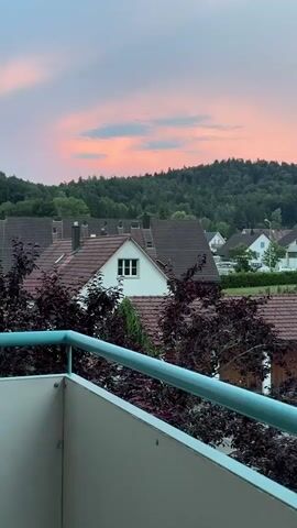 The video shows a view from a balcony overlooking a village at dusk. The sky is a soft pink and purple, with hints of blue. In the foreground, there is a balcony railing with a blue bar. Below the balcony, there are several houses with red tiled roofs and dark green trees. In the background, a dense forest covers a hillside. A silver car is parked in front of a garage on the right side of the screen. The overall impression is one of a peaceful, rural evening.