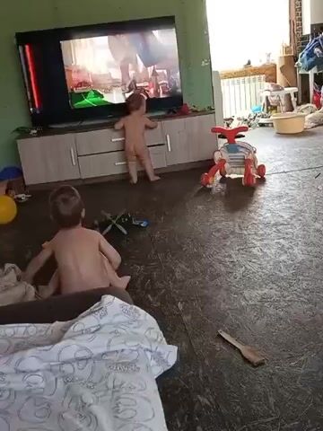 Two young boys, who appear to be siblings, are playing in a room. The older boy is sitting on the floor, while the younger boy is standing and holding onto a TV stand, watching television. Both children are nude. The TV is displaying an animated program. The room has dark flooring, and the walls are painted a light green. Various toys and household items are scattered around the room, including two yellow balloons, a red and white ride-on toy, and a small toy helicopter. In the foreground, part of a bed with a white and gray patterned duvet is visible. The scene appears to be indoors, possibly in a living room, with natural light entering from a window on the right side of the frame. Based on the timestamp, the video was captured in the afternoon.