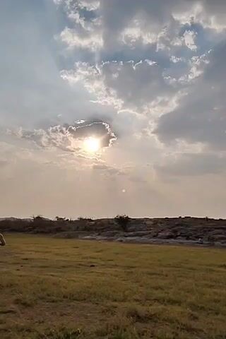 The video begins with a low-angle shot of a sunny sky with some clouds, the sun partially obscured. Below, there is a grassy field with a ridge of rocky land in the mid-ground. A voice can be heard, possibly calling out. The camera pans upwards, then to the right, revealing a clear sky and a large body of water in the distance. A pole, possibly a street lamp, stands in the foreground, with a wire stretching across the scene. In the distance, there are landforms which appear to be hills or mountains. The scene has a calm and peaceful atmosphere.