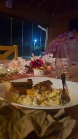 The video shows a close-up, table-level view of a messy dining table after a meal. Plates with leftover food and crumbs are visible, along with used cutlery. Empty glasses and a half-eaten cake suggest a celebratory occasion, possibly a party, has just concluded. Decorative elements, such as pink and gold balloons and flowers, are scattered across the table, reinforcing this impression. The scene is set indoors within a wooden-paneled structure, with a large glass window in the background revealing a dark, outdoor environment illuminated by distant lights. The overall impression is one of a lively gathering that has just ended, leaving behind the remnants of food and celebration.