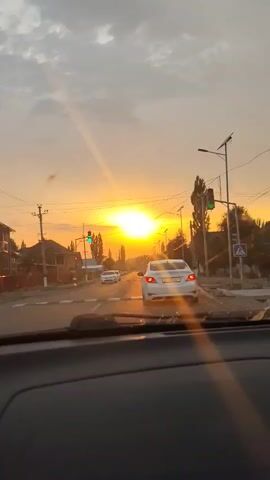 This is a first-person perspective video taken from inside a car, looking out through the windshield. The scene is a street in what appears to be a village or small town in Kara-Kochkor, Kyrgyzstan. The time of day is late afternoon or early evening, as evidenced by the bright, low-hanging sun casting long rays across the scene. The sky is a mix of cloudy and clear, with the sun creating a strong golden hue.

The main subject is the road ahead, with several cars visible. In the foreground, the dashboard of the car is partially visible. A white sedan is directly ahead, in the same lane. Further down the road, two other white cars are also visible. There are traffic lights at an intersection, with one showing green and another showing red. Telephone poles and overhead wires are a prominent feature of the setting, along with houses and trees lining the street. The overall impression is one of a typical evening commute or journey through a rural or semi-urban area.

Notable details include the vibrant golden light of the setting sun, which creates lens flares and a warm atmosphere. The presence of traffic lights and the road markings suggest a functioning road network. A street sign with a pedestrian symbol is visible on the right side of the road. The license plate of the white car in front is partially visible and appears to have Cyrillic characters, consistent with a Central Asian location like Kyrgyzstan. The weather appears to be clear with some clouds. The mood is somewhat serene and picturesque due to the sunset.