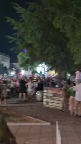 The video shows a crowded outdoor scene at night, likely a festival or outdoor event. In the foreground, several tables are set up with people seated and dining. The tables are adorned with various items such as drinks, food, and what appear to be cigarettes. The people in the foreground are engaged in conversation and eating. Behind them, a large gathering of people are visible, some standing and others dancing. A stage or performance area is set up in the background, illuminated by colorful lights, including purple and white lights. There are large green trees bordering the area, and buildings are visible in the distance. The overall atmosphere appears lively and festive.