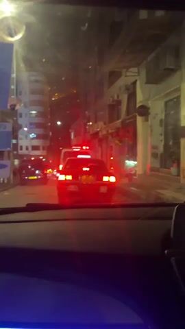 The video is a first-person view from inside a car, looking out through the windshield at a narrow street at night. The street is illuminated by streetlights and the taillights of several vehicles in front of the camera car. A taxi is directly ahead, and its brake lights are brightly illuminated. A white van is visible behind the taxi. On either side of the street, buildings line the road, with some shops and apartments visible. The buildings are decorated with some festive red decorations. The camera is stationary, as the car is stopped in traffic. The overall atmosphere is urban and somewhat congested.