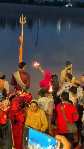The video shows a group of people gathered near a body of water, likely for a religious ceremony. In the foreground, a person dressed in a maroon and gold sash holds a flaming object, possibly a diya, towards a woman in a pink headscarf who is holding her hands together in prayer. Another person, holding a child, is positioned behind the woman. A trident symbol, possibly associated with Hindu deities, stands tall behind the person in the sash. The background features the dark water with reflections of lights. The overall atmosphere appears to be one of reverence and community participation.