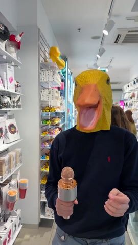 The video shows a person in a store wearing a yellow duck mask and a navy blue sweater with a small red logo. The person is holding a gold microphone in their right hand. They point their left index finger upward and move the microphone around as if singing or speaking into it. The store shelves are filled with various items, including hats, gift boxes, and decorative items. Other shoppers are visible in the background. The store has bright, even lighting.