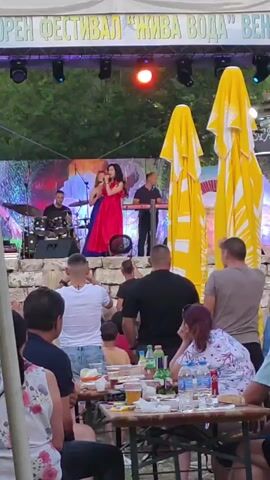 This video captures an outdoor music festival with a live band performing on a stage. The stage is set up with a backdrop and lighting. A female singer in a red dress is at the front of the stage, while other band members play instruments like drums and keyboards. A crowd of people is gathered in front of the stage, some standing and some seated at tables. In the foreground, there are tables with food and drinks, suggesting a relaxed, festive atmosphere. Large yellow umbrellas are visible on the right side of the frame. The event appears to be taking place in the late afternoon or early evening, with soft lighting. The backdrop of the stage has text that appears to be in Cyrillic, and the overall setting suggests a community gathering or cultural event. The inscription visible on the banner above the stage reads "МУЛТИКЛУРЕН ФЕСТИВАЛ \"ЖИВА ВОДА\" ВЕНЕЦА".