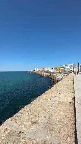 A high-angle, vertical, wide shot shows a bright blue sky over a calm, deep blue ocean with turquoise hues. The ocean meets a curved stone embankment or seawall that runs diagonally from the bottom left to the middle right of the frame. The seawall is constructed of large, light brown, rough-hewn stone blocks. Beyond the seawall, a rocky breakwater extends into the sea, dotted with a few scattered rocks and some vegetation. Along the shore behind the breakwater, a row of colorful buildings, painted in yellow, orange, green, and white, stand against the sky. A white building with a few windows is visible in the distance to the left, and a white tower or structure is located further down the coastline. A paved walkway or path runs alongside the seawall in the foreground, with a few people visible walking on it in the far distance on the right. The sun is shining brightly, creating a clear, distinct shadow on the seawall in the foreground. The water is smooth with very slight ripples.