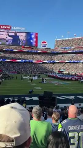 Here is a description of the video:
The video shows a large crowd at Levi's Stadium, presumably for a football game. People are standing while a large American flag is being unfolded on the field. A large screen above shows a man in front of the flag. There are also Pepsi and Levi’s Stadium advertisements visible. People are wearing various jerseys and clothing. The video appears to be shot from the stands. The sky is clear and bright.