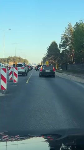 The video shows a view from a car stuck in traffic on a sunny day. Cars are lined up on the road ahead, with a yellow line separating lanes. On the right, there are trees lining the road. Streetlights are visible along the road as well. Red and white striped barriers are to the left of the car. The sky is blue. The dashboard of the car filming is visible.