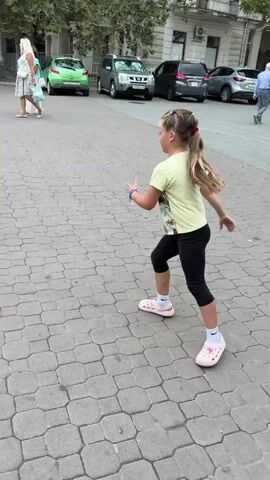 A young girl with blonde hair in a ponytail, wearing a light green t-shirt and black leggings, performs a series of athletic moves on a paved walkway. She is seen in profile, facing to the right of the frame. In the first frame, she is in a running stance. In the second frame, she is mid-air, with one leg bent and raised, in a jumping motion. The third frame captures her upside down, performing a cartwheel with her hands on the ground. She then transitions to a standing pose in the fourth frame, facing forward with her arms slightly outstretched. Finally, in the fifth frame, she returns to a pose similar to the first, with her body slightly turned and her arm extended. In the background, other people are walking, and cars are parked along the street. The environment appears to be an outdoor urban setting.