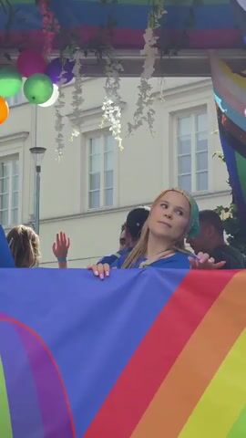 A young woman with blonde hair wearing a green bandana and a blue t-shirt is on a float. She is holding a rainbow flag. There are balloons and greenery around her. She is looking to the side and her mouth is slightly open. Another person, partially visible, is on the left holding a drink. In the background, buildings and trees are visible.