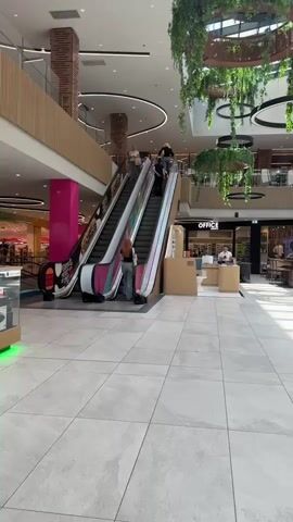 This video shows the interior of a modern, multi-level shopping mall. The floors are tiled with a light gray stone pattern, and the ceiling features a geometric design with recessed lighting and large circular frames adorned with hanging plants. Natural light streams in from a skylight. Escalators are visible on the left side of the frame, with people using them. Several shops and restaurants are visible on the upper levels and at ground level, including "OFFICE," "Jeordie's," and "CALZEDONIA." Some tables and chairs are set up outside one of the establishments, suggesting a seating area. People are seen walking and interacting within the mall. Sunlight casts distinct shadow patterns on the floor.