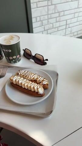 A close-up video shows two eclairs on a white plate, topped with toasted meringue, placed on a silver tray. Next to the plate are two coffee cups, one with black coffee and the other with latte art. A pair of sunglasses rests on the white table behind the cups. The background includes a tiled wall and a dark partition. The scene is well-lit, with a focus on the food and drinks.