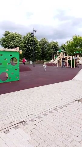 This video captures a playful scene at a playground in Orekhovo-Zuyevo, Russia. The playground features a bright green climbing wall, a wooden swing set with blue seats, and a red rubberized surface. Several children are present, actively engaged in play. One child on a scooter zooms across the playground, while others are on the swings, enjoying the motion. In the background, lush green trees and a cloudy sky suggest it's daytime, likely overcast. The overall mood is one of carefree fun and activity. A park bench sits near the playground, with a pink bicycle and a red scooter parked beside it, indicating that visitors often arrive on these modes of transport. The surrounding area appears to be a park or recreational space, with a paved walkway made of light-colored bricks bordering the playground. The setting seems to be a typical suburban park, offering a space for children to play and families to relax.