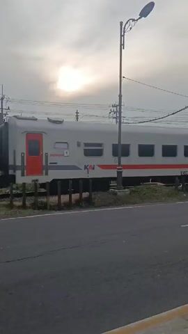 The video shows a train moving to the right on a track next to a road. The train cars are painted gray and white with a red stripe. The road appears to be asphalt and has yellow lines on the edge. There are trees lining the track and road. A billboard is briefly visible on the left side of the frame. The sky is overcast and the sun is shining through the clouds. The camera follows the train as it moves.