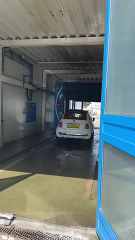A white Fiat 500 with a red roof and a Dutch license plate is slowly driving into a car wash. The car is positioned in the center of the frame as it enters the covered wash bay. The car wash entrance is framed by bright blue doors and glass panels, with the doors partially open. The car wash bay itself has a metal ceiling and walls, with visible pipes and equipment. Water is present on the floor of the car wash, indicating it is operational or has recently been used. The exterior shot shows the car wash structure with its blue entrance doors and a portion of the surrounding area, which appears to be an outdoor lot with paved ground marked with white lines. The weather seems to be sunny.