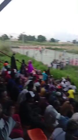 The video shows an outdoor gathering of people watching an event on a court. The camera is positioned above and behind a crowd of spectators seated in bleachers. The spectators are facing a fenced-off court area where a group of people are gathered. Some individuals in the court area appear to be interacting, possibly dancing or performing. The court is surfaced with red and green material and is surrounded by a chain-link fence. Beyond the court, there are grassy fields, trees, and some buildings in the distance. The sky is overcast. The video is shaky, suggesting it was recorded handheld.