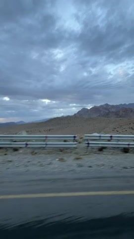 The video begins with a view of a desolate landscape of mountains and dry terrain. A road with a yellow line marking the edge is visible in the foreground, with a metal guardrail separating it from the rough terrain. The sky is overcast with heavy clouds, suggesting a cloudy or possibly stormy day. As the video progresses, the camera, which appears to be mounted on a moving vehicle, pans across the scene, revealing more of the mountainous terrain and the road stretching into the distance. A truck can be seen driving on the road ahead. The camera appears to be attached to a scooter as a handlebar and part of a mirror are visible in the lower frame.