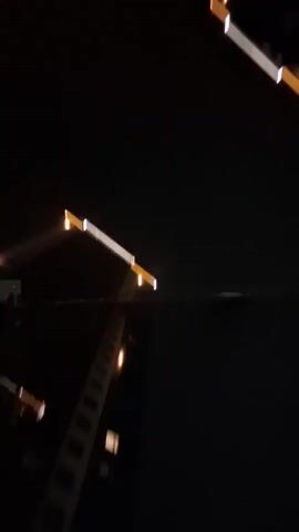 The video shows a night scene of buildings with lights. There are two large buildings visible, one in the foreground and another in the background on the right. The buildings are dark, but illuminated by lights. The building in the foreground appears to be a dark structure with lights along its edges, possibly outlining its shape or structure. There are also visible lights on what appears to be a ladder or a series of steps ascending the side of this building. The building in the background on the right is also dark with bright lights, including some blue lights, visible near its top. The camera perspective is looking upwards, towards the sky and the tops of the buildings. There are no people visible in the video. The scene is blurry, suggesting movement of the camera or the subject.