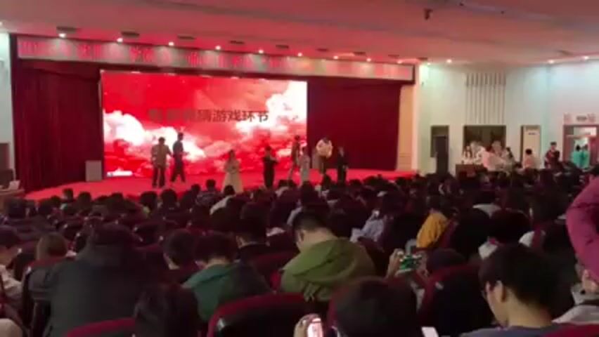 A group of young people is on a stage with a large audience in front of them. There are 4 men and 2 women on stage. The men are wearing casual clothing, and the women are wearing dresses. The stage is brightly lit with a large LED screen behind the performers. The screen displays a red graphic with Chinese writing. The audience is seated in rows of red chairs. Many audience members are holding up their phones, presumably recording the performance. The room is large and brightly lit.