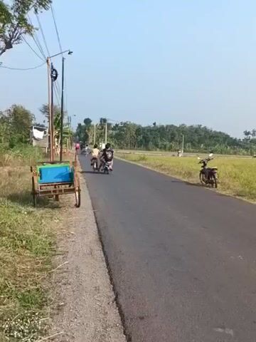 A two-wheeled motorized cart with a blue container is parked on the left side of a paved road. Further down the road, several motorcycles are visible. One motorcycle, ridden by a person wearing a yellow shirt, is moving away from the camera. To the right of the road, a field of dry grass and some trees are visible. The sky is clear and blue.