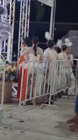 A group of people are on a stage. Some are wearing white outfits and others are in casual clothes. One person is speaking into a microphone. A band is playing instruments in the background. There are white metal barriers in front of the stage. The event appears to be outdoors at night. A sign on the stage has Thai writing and dates.