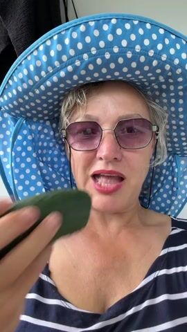 A woman is captured in a close-up, selfie-style shot, wearing a blue and white polka-dotted sun hat and sunglasses with pink lenses. She has a striped shirt on, and her hair is blonde and curly. She is holding a piece of cucumber up to her mouth and appears to be biting into it. The background is blurred, but hints of a white wall and dark fabric suggest an indoor or shaded outdoor setting. The lighting appears natural, indicating it could be daytime. The woman's expression is neutral as she chews. There is no visible text in the frame. The overall mood is casual and possibly summery.