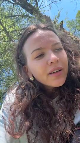 A young woman with long, curly brown hair and green eyes is looking at the camera. She is outdoors, with trees and a blue sky visible in the background. She is wearing a white t-shirt and large pearl earrings. She touches her face and mouth as she speaks.