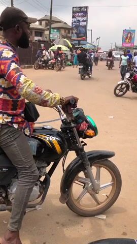 A man wearing a colorful patterned shirt and a baseball cap sits on a motorcycle. He is looking to his left. A person is walking by on his right side. In the background, other people are walking and riding motorcycles. There are buildings and billboards in the distance. The ground appears to be dirt and gravel. The video is taken from a side angle of the motorcycle.