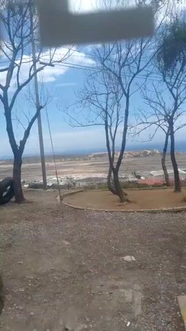The video displays an outdoor panoramic view, panning slowly from left to right. In the foreground, there is a dirt ground or road, with several trees, some bare and some with sparse foliage, framing the scene. Overhead, power lines are visible. The background reveals a wide landscape, featuring what appears to be an airport or an industrial area with various structures, extending towards a body of water, likely the sea or ocean, under a partly cloudy blue sky. The time of day is daylight. No people or text are visible in the video. The location context is Catia La Mar, Venezuela.